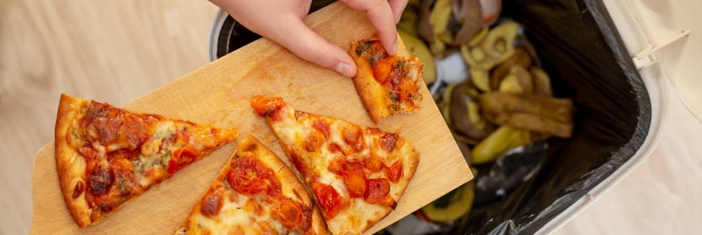 Pizza dilimleri tahtanın üzerinde; bir el artık yemek artığını çöp kutusuna atıyor — gıda israfı